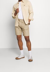 Beżowa koszula z guzikami na białym t-shircie, zestawiona z beżowymi szortami i białymi skarpetkami. Buty są jasne, z brązową gumową podeszwą.