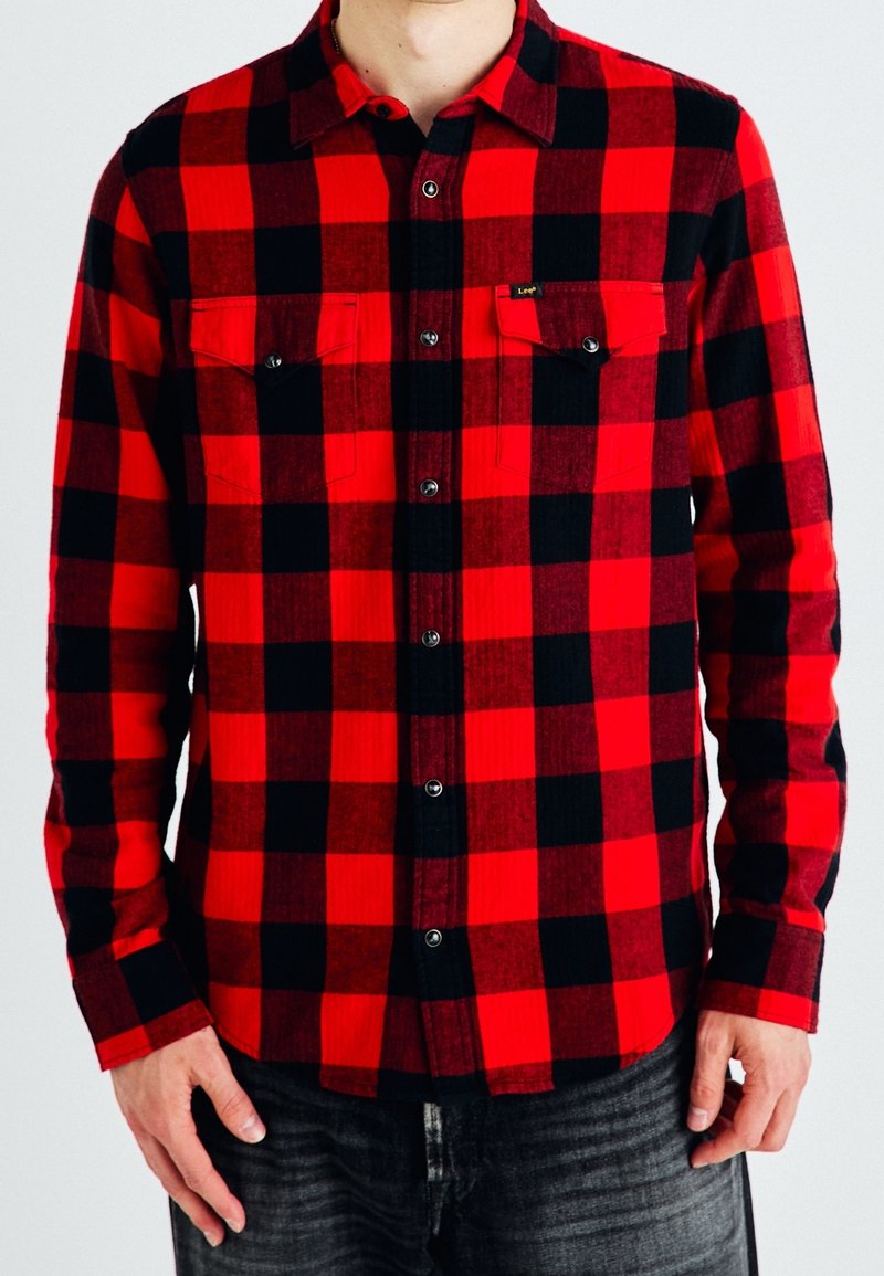 Homme portant une chemise en flanelle à carreaux rouge et noir avec des poches boutonnées et un jean foncé sur un fond clair uni.