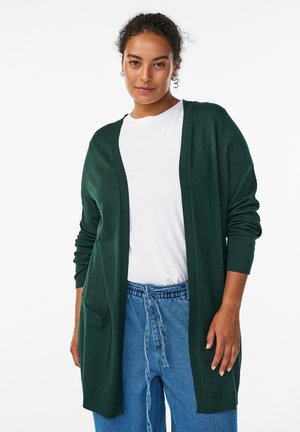 Femme portant un cardigan vert foncé ouvert sur un t-shirt blanc et un jean bleu large, debout devant un fond blanc uni.