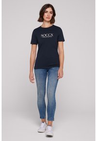 T-shirt en coton bleu marine avec le logo "SOCCX TIMELESS EDITION", manches courtes, associé à un jean skinny bleu clair et des baskets blanches.