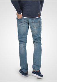 Persona con jeans de color azul claro y zapatillas azul marino, con una mano en el bolsillo trasero, de pie sobre un fondo blanco.