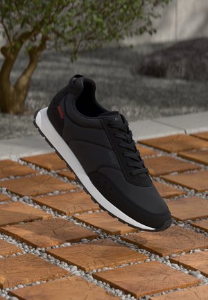 Czarny sportowy sneaker z białą podeszwą, czerwonym detalem i sznurowadłami, spoczywający na drewnianych kafelkach z małymi kamieniami na zewnątrz.