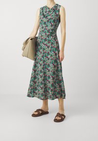 Vestido midi floral en verde con acentos en rosa y beige, escote drapeado y una silueta fluida. Combinado con sandalias tejidas marrones y un bolso beige.