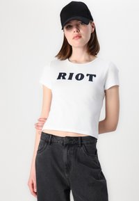 Bela skrajšana majica s kratkimi rokavi in črnim napisom "RIOT", v kombinaciji z visokim pasom črnimi kavbojkami iz teksturirane tkanine, preprostega dizajna.