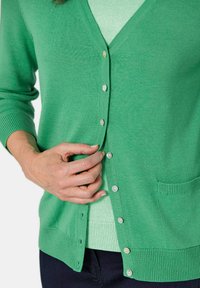 Grüne Strickjacke aus weichem Material, mit tiefem V-Ausschnitt, sechs Knopfverschlüssen und einer Seitentasche. Über einem hellgrünen Oberteil getragen.