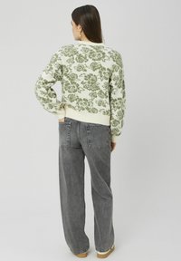 Femme aux longs cheveux bruns portant un pull blanc à motif floral vert et un jean gris ample, debout avec une main dans la poche arrière.