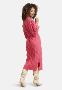 Abito lungo rosa lavorato a maglia con motivo a diamanti testurizzati, vita con cintura e spacco laterale. Abbinato a stivali crema al ginocchio.