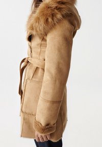 Tan-farvet faux ruskind frakke med hætte foret med pels, taljebælte og pelskant ved ærmekanterne. Har lynlåslukning og lommedetaljer.