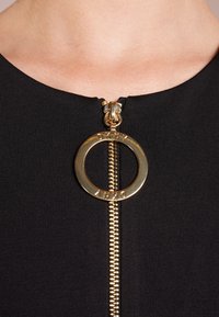 Gros plan sur la tirette dorée DKNY sur un vêtement en tissu noir au niveau du cou d'une personne.