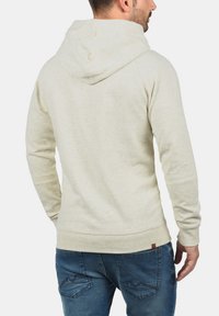 Light beige hooded sweatshirt made of textured fabric. It features long sleeves and a kangaroo pocket. The hem has a small brand tag.