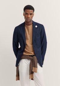 Blazer navy con una finitura testurizzata, maglione marrone e sciarpa di maglia a righe annodata in vita, abbinata a pantaloni bianchi.
