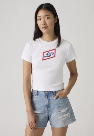 Mujer con cabello largo y oscuro, vistiendo una camiseta blanca de Levi's y unos pantalones cortos de mezclilla azul claro con rasgaduras, de pie con una mano en el bolsillo.