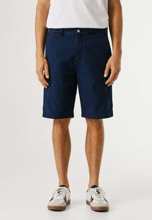 Hombre vistiendo pantalones cortos cargo azul marino, zapatillas blancas y una camisa blanca, de pie contra un fondo claro y liso.