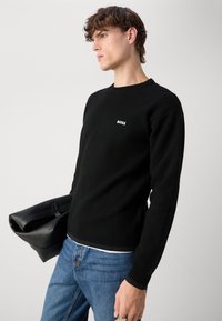 Czarny dziany sweter z prążkowanymi mankietami i dołem, z małym białym logo "BOSS" na piersi. Noszony z niebieskimi dżinsami i czarną torbą.