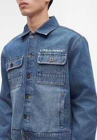 Άνδρας που φοράει μπλε τζιν μπουφάν με κουμπιά και δύο τσέπες στο στήθος, η μία με άσπρο κείμενο που γράφει "I./A/B c/o Off-White".