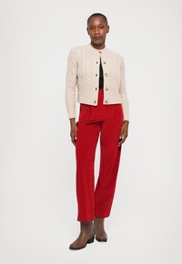 Light beige knitted cardigan with cable pattern, gold buttons, paired with loose red corduroy pants and brown ankle boots.