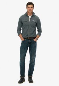 Man in een donkergrijze button-up shirt met opgerichte kraag, blauwe jeans, zwarte schoenen, een bruine riem en een polshorloge staat met zijn handen in zijn zakken.