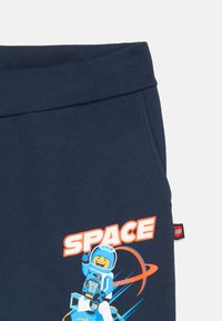 Dunkelblaue Hosen mit geripptem Bund, versehen mit einer lächelnden LEGO-Astronautenfigur, orangefarbenen Ringen und dem Wort "SPACE" in fett gedruckten weißen und orangeschwarzen Buchstaben.