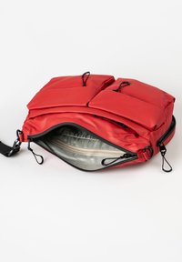 Red nylon bag with two front pockets, zipper closure, and a silver interior. It features adjustable straps and pull tabs.
