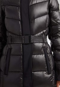 Chaqueta puffer negra con un acabado brillante, diseño acolchado, cinturilla elástica y cierre de hebilla. Se pueden ver los bolsillos laterales.