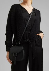 Cárdigan negro de punto con frente abierto y cierre de lazo sobre una camisa negra de botones. Pequeño bolso cruzado de cuero negro con detalle de borlas.