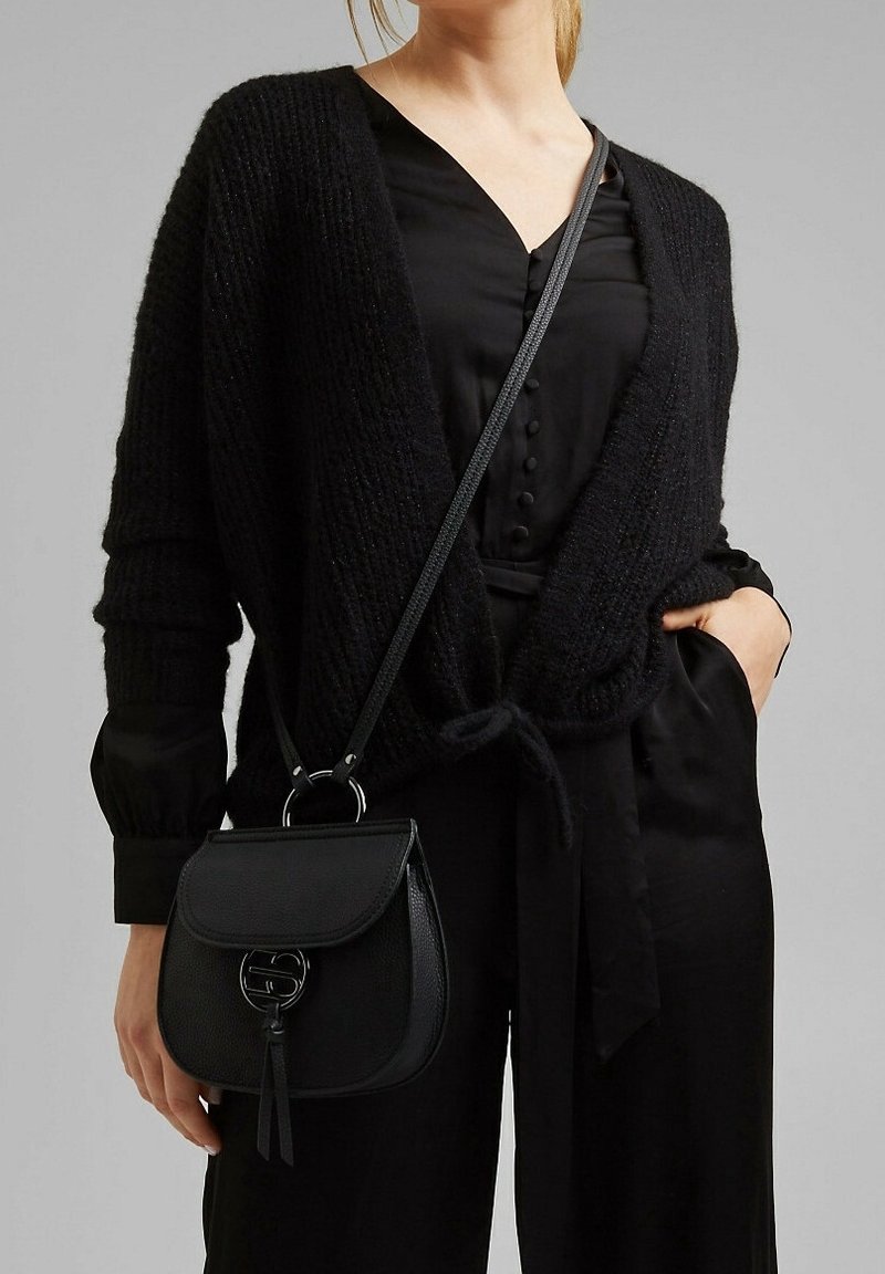 Cárdigan negro de punto con frente abierto y cierre de lazo sobre una camisa negra de botones. Pequeño bolso cruzado de cuero negro con detalle de borlas.