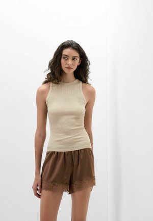 Vrouw met lang donker krullend haar, gekleed in een beige mouwloze top en bruine short met kanten rand, staand tegen een effen lichte achtergrond.