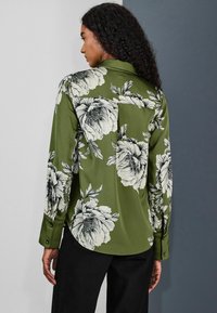 Zelená kvetinová blúzka s dlhými rukávmi, zdobená veľkými bielymi kvetinovými vzormi, klasickým golierom a detailmi s gombíkmi na manžetách.