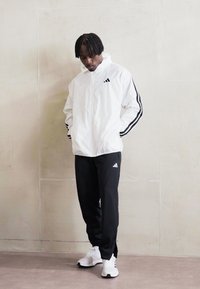 Casaco corta-vento branco com riscas pretas, calças de jogging pretas e sapatilhas brancas, com detalhes de marca em cada peça.