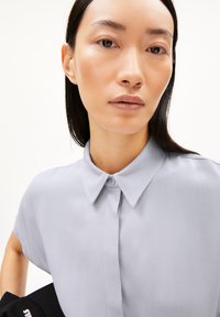 Lichtgrijs kortvrij shirt met een spread collar, knoopsluiting en een gladde textuur, gedragen met een zwart accessoire op de schouder.