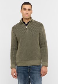 Olivgrüner, gestrickter Pullover mit vertikalem Rippmuster, halblangem Reißverschluss am Kragen und tailliertem Bund. Kombiniert mit blauen Jeans.