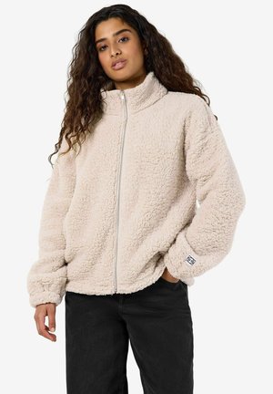 Kvinde med langt krøllet hår iført en lys beige fleecejakke med lynlås og sorte bukser, står med den ene hånd i lommen.