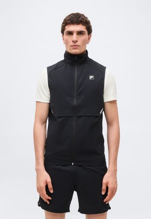 Homme portant un gilet sans manches noir à fermeture éclair sur une chemise blanche à manches courtes et un short noir, debout devant un fond clair uni.