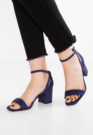 Pieds portant des sandales à talons blocs bleu marine avec brides à la cheville, associées à un pantalon court noir à ourlet effiloché sur un sol blanc.