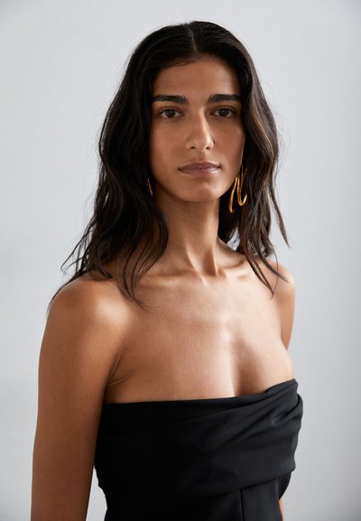 Schwarzes trägerloses Kleid mit gerafften Stoffdetails und glatter Textur. Das Model hat lange, wellige, dunkle Haare und trägt große goldene Creolen.