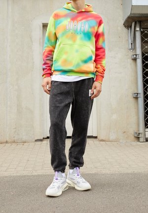 Osoba nosząca wielokolorową bluzę z kieszenią kangurka Jordan Nike w stylu tie-dye, czarne spodnie dresowe i białe trampki, stojąca na brukowanej powierzchni na zewnątrz.
