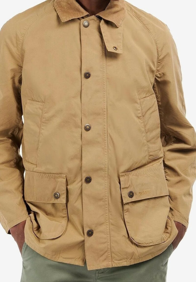 Veste beige avec col en velours côtelé, fermeture à boutons et quatre poches, portée sur un pantalon vert, mains partiellement dans les poches.