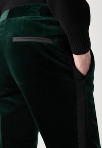 Green velvet trousers with a smooth texture, featuring a black side stripe and a back pocket detail. The waistband is flat and simple.