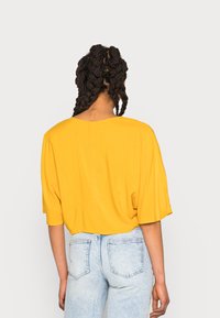 Top amarelo de mangas curtas com um corte descontraído e decote redondo, combinado com jeans de cintura alta de tom azul claro, exibindo uma textura de tecido suave.