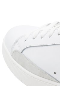 Sneaker bianco con tomaia in pelle liscia, accento in camoscio grigio a contrasto, lacci bianchi e suola in gomma testurizzata. Dettaglio di cucitura visibile.