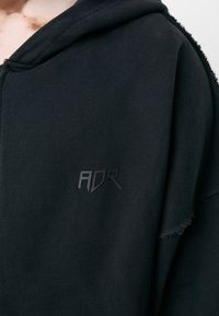 Black hoodie with a smooth texture, featuring a zip-up design and a distressed hem. It has a prominent black "ADR" logo.