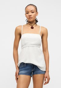 Γυναίκα με πλεξούδες, που φοράει λευκό αμάνικο τοπ, μπλε τζιν σορτς και μαύρο κολιέ με κόκκινο μενταγιόν σε σχήμα λουλουδιού.