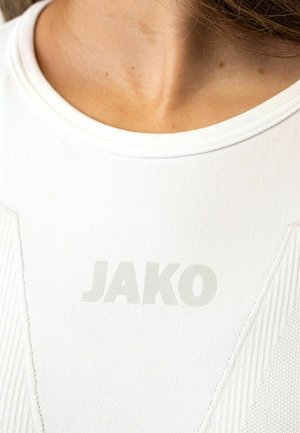 Primo piano di collo e parte superiore del petto che indossano una maglia sportiva bianca con trama, con il logo "JAKO" stampato in modo sfocato sulla parte anteriore.