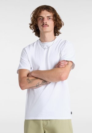 Weißes T-Shirt aus Baumwolle mit kurzen Ärmeln und geradem Schnitt. Auffällige Merkmale sind ein kleines Logo am Saum und sichtbare Tattoos an den Armen.