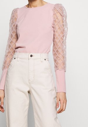 Mujer con una camiseta rosa claro con mangas abullonadas transparentes con textura y pantalones crema de talle alto sobre un fondo liso.