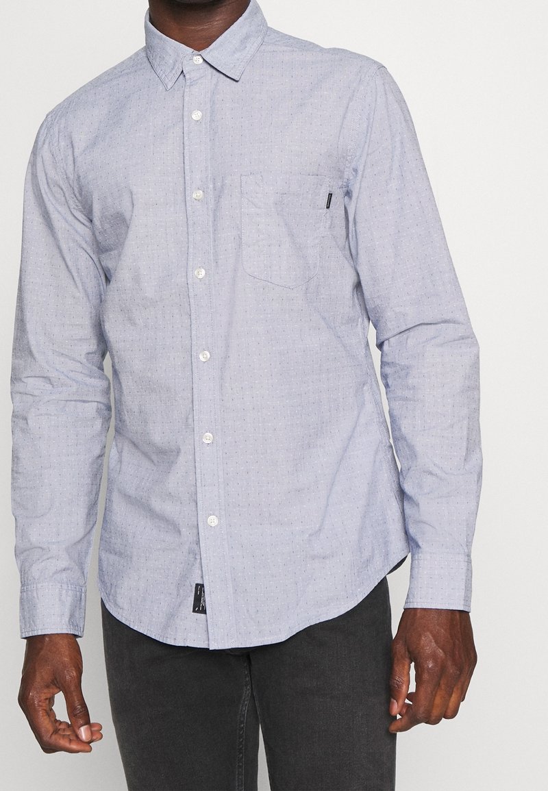 Homme portant une chemise à manches longues bleu clair avec un motif discret à pois et une poche poitrine, associée à un pantalon foncé.