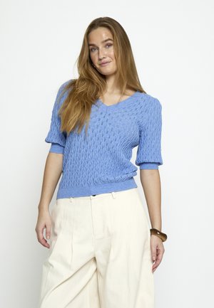 Žena s dlhou vlasmi, oblečená v modrom textúrovanom svetri s krátkym rukávom a voľných bielych nohaviciach na jednoduchom bielom pozadí.