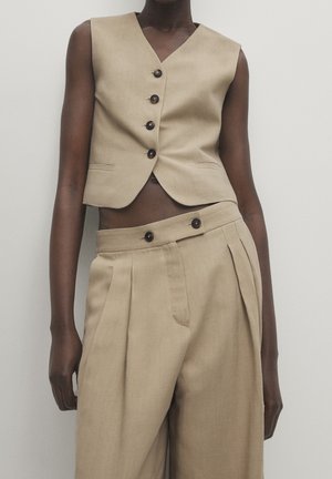 Osoba o ciemnej karnacji ubrana w beżową, bezrękawnikową kamizelkę zapinaną na guziki oraz pasujące wysokiej talii spodnie z zakładkami, stojąca na tle jednolitego tła.