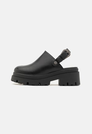 Sabot en cuir noir avec sangle ajustable et semelle en caoutchouc épaisse, présentant une finition texturée et un détail de boucle sur le côté.