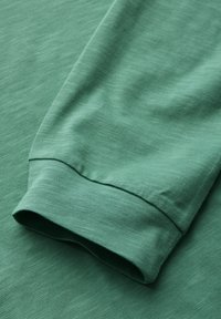 Lähikuva vihreast pitkähihaisesta paidan hihasta, jossa näkyy ompelu ja pehmeä, tekstuurinen kangas.